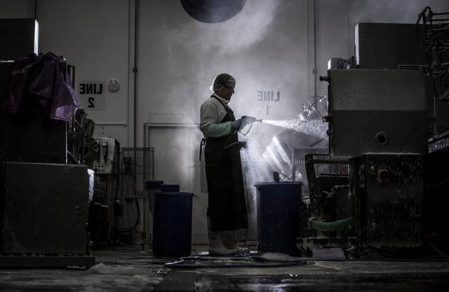 Man wearing protective clothing spraying industrial equipment clean, in a dark and steam-filled environment
